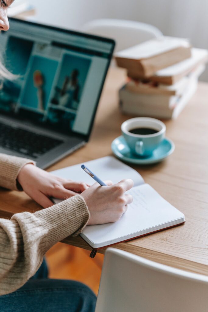 A thoughtful individual reading a book at a desk with a laptop and notepad nearby.